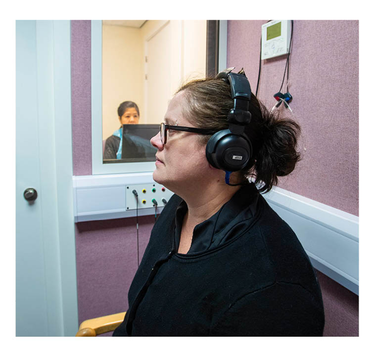 Patient wearing headphones in soundproof room, with audiologist in room outside. Audiologist can be seen through the window.