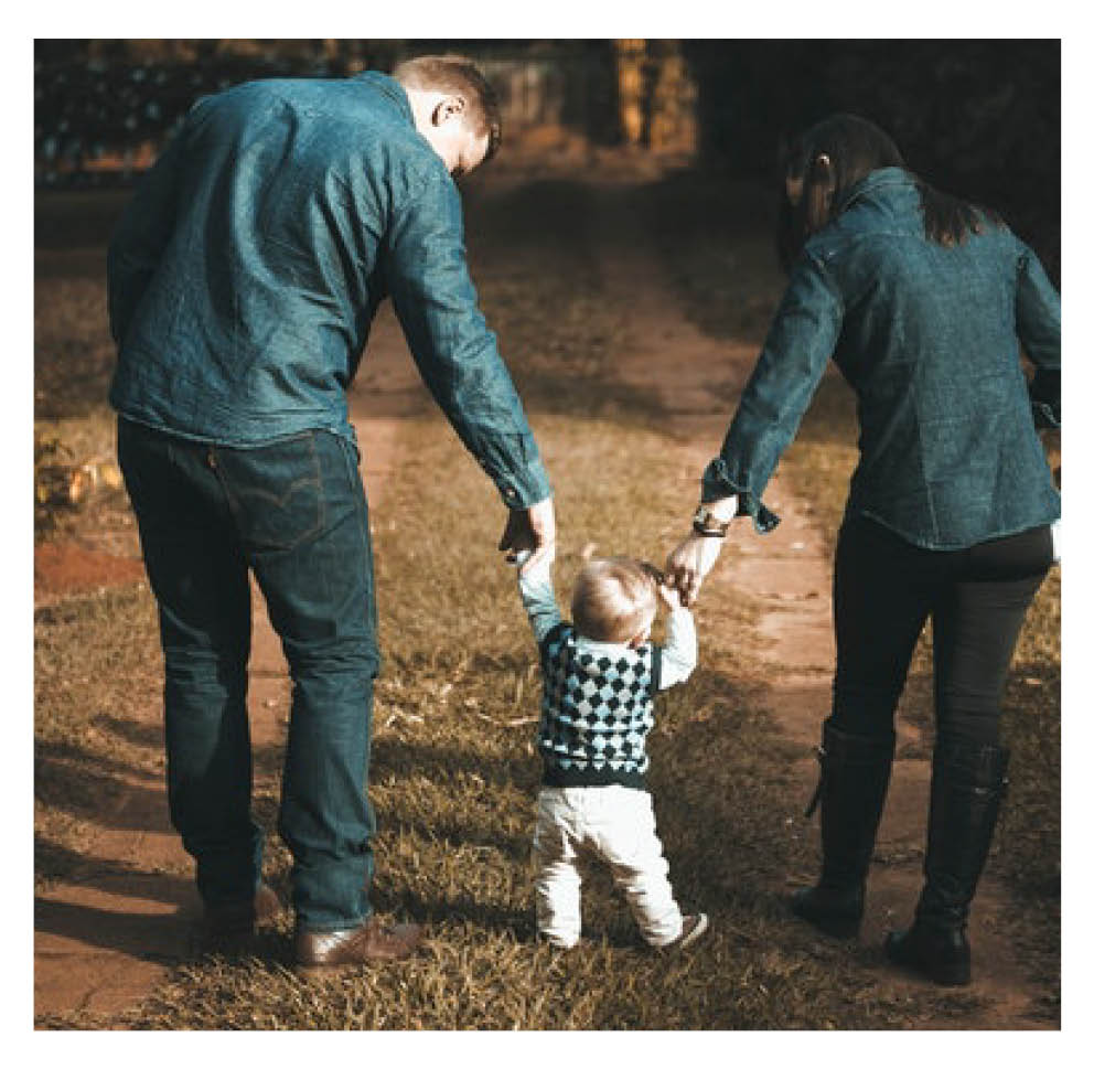 Parents each holding one of their child's hands, supporting them while they learn to walk.